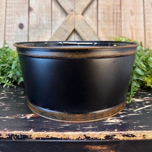 May include: Black metal planter with a distressed gold rim. The planter is round and has a wide opening. It is sitting on a dark wooden surface.