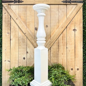 May include: A tall, white candle holder featuring a square base that transitions into a rounded, turned-style column with a circular top platform. The object has a smooth, matte finish and stands upright as a singular, solid decorative piece.