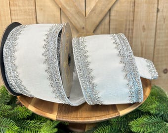 May include: A spool of white ribbon featuring a metallic silver decorative trim along both edges. The ribbon has a smooth, solid white center and a textured, scalloped silver border that runs the entire length of the fabric on both sides.