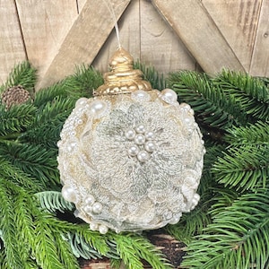 Pu&ograve; includere: Una decorazione natalizia sferica caratterizzata da un rivestimento in pizzo color crema con motivi floreali, arricchita da grappoli di perle artificiali bianche e un tappo in oro metallizzato, sospesa tramite un filo trasparente.