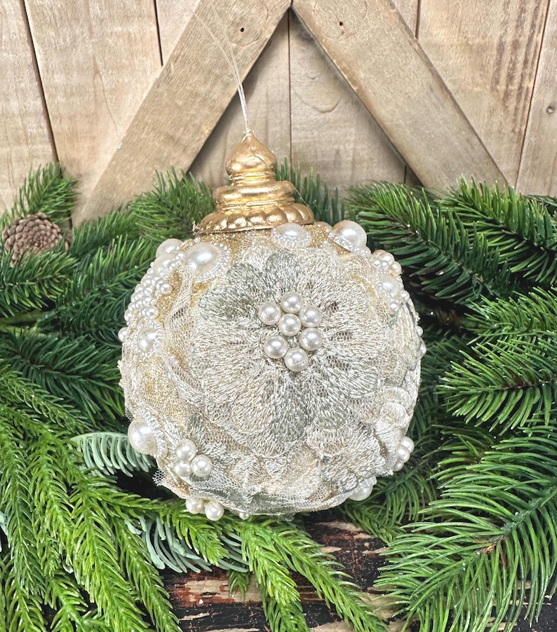 Pu&ograve; includere: Una decorazione natalizia sferica caratterizzata da un rivestimento in pizzo color crema con motivi floreali, arricchita da grappoli di perle artificiali bianche e un tappo in oro metallizzato, sospesa tramite un filo trasparente.