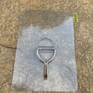 May include: A clear plastic adhesive hook with a silver metal hook. The hook is attached to a clear plastic backing with the word "OPEN" printed on a yellow sticker.