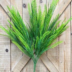 May include: A vibrant green artificial grass plant with long, slender leaves and a full, textured appearance. The plant has a bundle of stems and is set against a rustic wooden background, creating a natural aesthetic. Ideal for home decor.