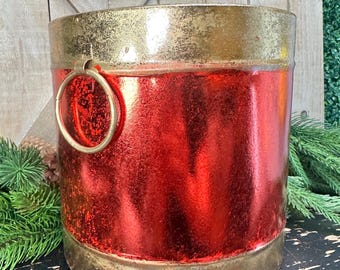 May include: A cylindrical metal bucket featuring a vibrant red centre band with a distressed finish, contrasted by a metallic gold rim and base. The container includes a circular gold ring handle attached to the side and rests on small feet.