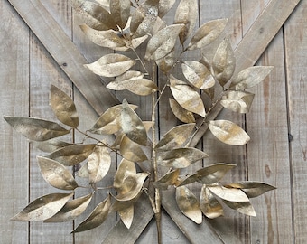 May include: A decorative artificial branch featuring numerous oval-shaped leaves with a metallic gold finish. The leaves have a textured, speckled appearance and are arranged on a central stem that branches out into multiple smaller stems.