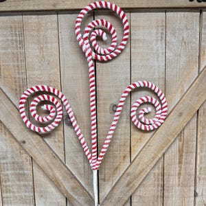 May include: A decorative Christmas stem with a red and white striped design. The stem features three swirling spiral shapes and a white base, set against a wooden background. The festive design is reminiscent of candy canes.
