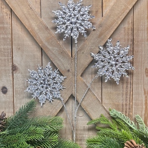 May include: Three silver glitter snowflake ornaments on stems, arranged against a rustic wooden door. The snowflakes are detailed and sparkling, with a natural evergreen and pinecone base. Perfect for holiday decor.