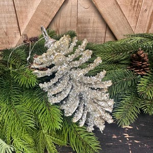 May include: A silver and crystal-embellished pinecone ornament rests on a bed of green pine needles and small pinecones. The ornament has a silver metal loop for hanging. The background is a wooden surface.