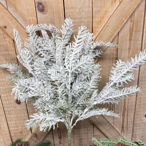 May include: A faux frosted evergreen branch with green needles and a dusting of white. The artificial plant has a central stem and is surrounded by other evergreen branches and a pinecone, all against a wooden background.