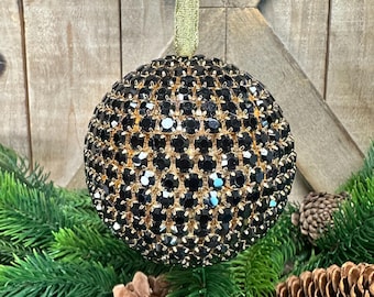 Può includere: Un ornamento sferico ricoperto di strass neri sfaccettati incastonati in una montatura in metallo dorato. L'ornamento è sospeso da un sottile nastro dorato scintillante fissato sulla parte superiore, creando una superficie dall'aspetto riflettente e strutturato.