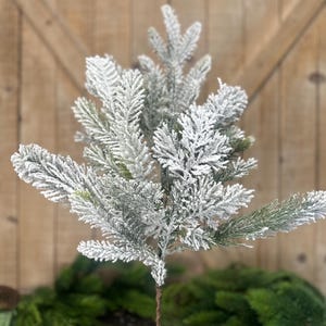 May include: A decorative artificial evergreen branch featuring dense, needle-like foliage coated in a thick layer of white, snow-like flocking. The branch has a central brown, twisted wire stem that supports multiple smaller, textured green sprigs.