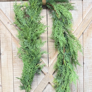 May include: Two green artificial cedar garlands hanging against a light brown wooden door. The garlands are made of faux cedar branches and are attached to the door with a brown leather loop. Decorative black metal hinges are visible.