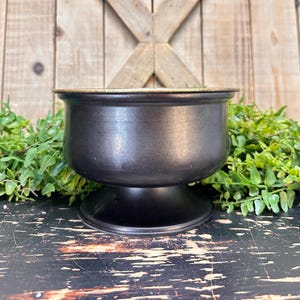 May include: Black metal planter with a gold rim and a pedestal base. The planter is sitting on a wooden surface.