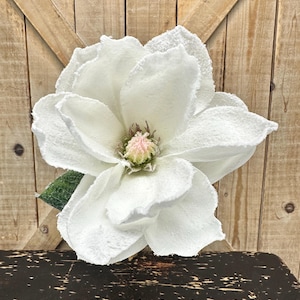 May include: A large, artificial white magnolia flower with a textured, frosted appearance on its petals. The center of the bloom features a pale pink and green stamen cluster. A single green leaf is visible tucked behind the lower left petals.