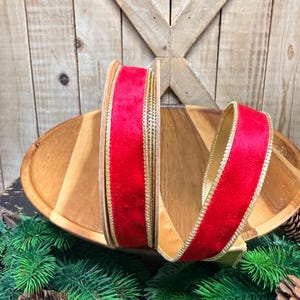 May include: A spool of red velvet ribbon featuring metallic gold edges. The ribbon is wound on a circular core, with a portion of the fabric extending outward to display the texture and the gold trim on both sides of the vibrant red material.