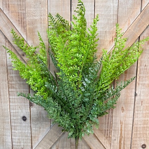 May include: A decorative artificial fern plant featuring multiple stems with finely detailed, serrated leaves in varying shades of vibrant lime green and deep forest green, arranged in a dense, upright cluster on a single central stalk.