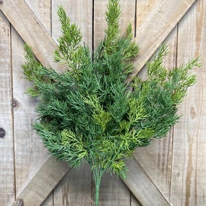 May include: A green artificial plant with multiple stems and a full, textured appearance. The plant has a variety of green shades, from light to dark, and is set against a wooden background. The plant is a decorative item.