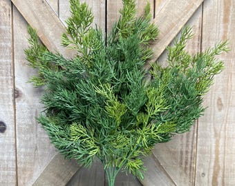 May include: A green artificial plant with multiple stems and a full, textured appearance. The plant has a variety of green shades, from light to dark, and is set against a wooden background. The plant is a decorative item.
