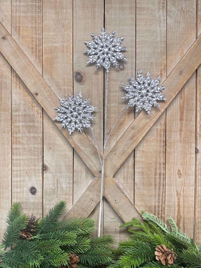 May include: Three silver glitter snowflake ornaments on stems, arranged against a wooden background. The snowflakes are detailed and sparkling, with a green garland and pinecones at the bottom. Perfect for holiday decor.