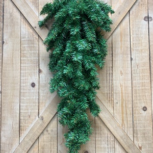 May include: A green artificial teardrop swag, made of faux evergreen branches, hangs against a rustic wooden door. The swag is a decorative accent for the home, often used for seasonal or holiday decor.