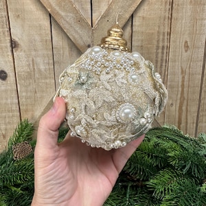 Pu&ograve; includere: Un ornamento sferico ricoperto di tessuto in pizzo color oro con un intricato motivo floreale. La superficie &egrave; impreziosita da numerose perle artificiali bianche di varie dimensioni e da un tappo in metallo dorato sulla parte superiore.