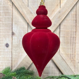 May include: A deep red, velvet-textured finial-shaped Christmas ornament with a rounded, tiered top and a pointed base. The ornament features a gold-colored metal cap at the top, designed for hanging, and has a smooth, matte, flocked finish.