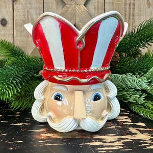 May include: A ceramic nutcracker head planter featuring a tall red and white striped hat with gold trim. The nutcracker has a pale face, blue eyes, a prominent nose, and a white mustache. The top of the hat is open, creating a hollow vessel.