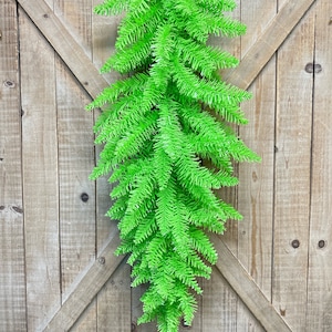 May include: A vibrant green artificial fir garland hanging against a rustic wooden door. The garland is made of dense, needle-like foliage, creating a full and textured appearance. A brown leather loop is used for hanging. This decorative piece adds a pop of color.