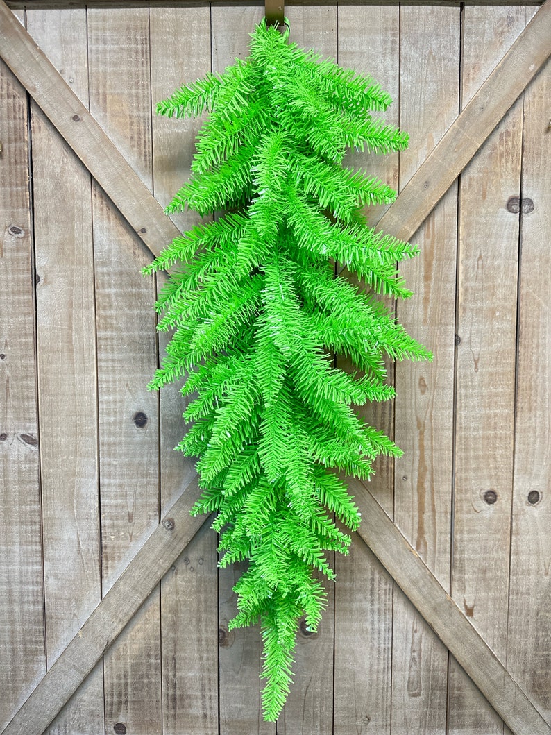 May include: A vibrant green artificial fir garland hanging against a rustic wooden door. The garland is made of dense, needle-like foliage, creating a full and textured appearance. A brown leather loop is used for hanging. This decorative piece adds a pop of color.