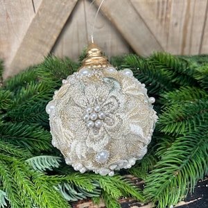 Pu&ograve; includere: Una pallina di Natale sferica con una base glitterata dorata ricoperta da un intricato ricamo in pizzo floreale color crema. L'ornamento &egrave; impreziosito da piccole perle bianche artificiali sparse sulla superficie e sormontato da un tappo dorato.