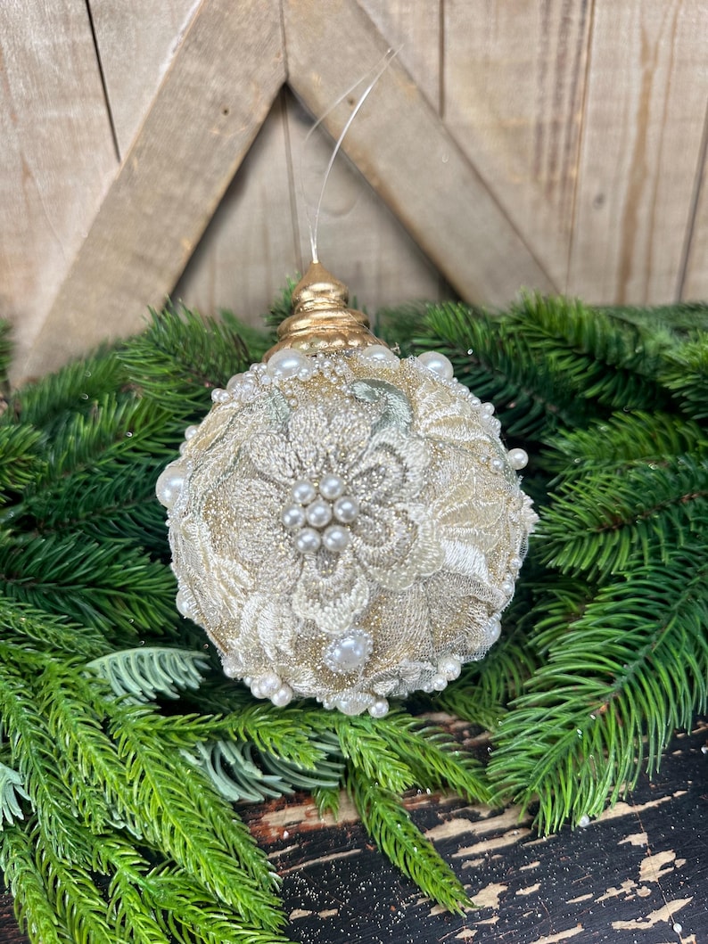 Pu&ograve; includere: Una pallina di Natale sferica con una base glitterata dorata ricoperta da un intricato ricamo in pizzo floreale color crema. L'ornamento &egrave; impreziosito da piccole perle bianche artificiali sparse sulla superficie e sormontato da un tappo dorato.