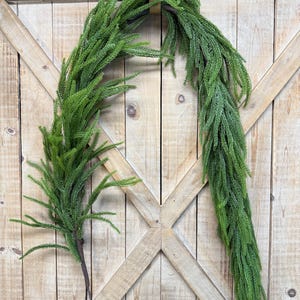 May include: A green artificial garland with a dark brown metal loop for hanging. The garland is made of small, needle-like leaves and is displayed against a light brown wooden door with decorative black hinges.