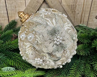 May include: A spherical Christmas ornament covered in gold lace fabric with intricate floral embroidery. The surface is embellished with numerous white faux pearls of varying sizes. A metallic gold cap sits at the top with a thin wire loop for hanging.