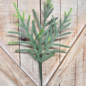 May include: A single artificial evergreen stem with a dark green stem and light green and dark green foliage. The faux plant is set against a light brown wooden background with a diagonal pattern.