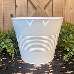 May include: White metal planter with a ribbed design. The planter is empty and sitting on a dark wooden surface.