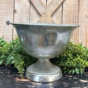 May include: A silver metal bowl with a pedestal base. The bowl has a distressed finish and is perfect for displaying flowers or other decorative items.