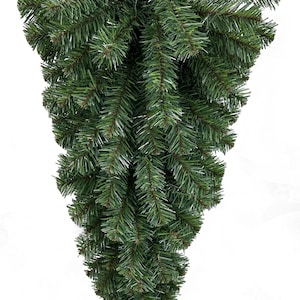 May include: A cascading, artificial Christmas swag composed of dense, dark green pine needles. The swag is designed to hang, with a full, textured appearance. The needles are detailed, creating a realistic look for holiday decor.