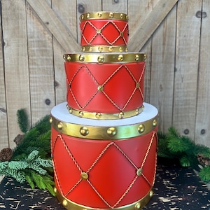 May include: A three-tiered decorative drum set featuring a vibrant red finish with gold-toned metallic trim. Each cylindrical drum is adorned with a raised gold diamond-shaped rope pattern and gold studs along the top and bottom edges.