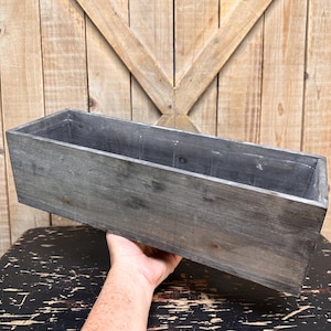 May include: A rectangular wooden planter box with a weathered gray finish. The box is held by a person's hand, showcasing its length and depth. The background features a wooden wall with a decorative pattern.
