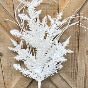 May include: A white, artificial fern spray with delicate, feathery leaves and thin, wispy stems. The faux plant has a textured, frosted appearance and is set against a rustic, wooden background. This decorative piece is suitable for floral arrangements and home decor.