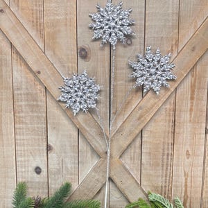May include: Three silver snowflake ornaments on stems, arranged against a wooden background. The snowflakes have a glittery texture and are accented with clear crystals. Green pine branches and pinecones are at the bottom.