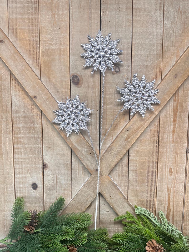 May include: Three silver snowflake ornaments on stems, arranged against a wooden background. The snowflakes have a glittery texture and are accented with clear crystals. Green pine branches and pinecones are at the bottom.