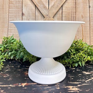 May include: White ceramic pedestal bowl with a fluted base. The bowl is round and has a wide opening. It is sitting on a dark wooden surface.