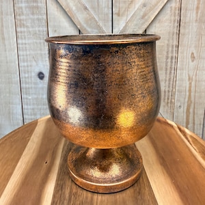 May include: A bronze-colored, pedestal planter with a wide, rounded body and a flared rim. The metallic finish has a textured appearance with darker accents, creating a vintage aesthetic. The planter sits on a round, pedestal base.