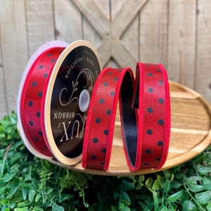 May include: A spool of red ribbon with black polka dots. The ribbon is labeled "LUX RIBBON VALUE SELECTION". The ribbon is displayed on a wooden tray with greenery in the foreground. The background is a wooden door.