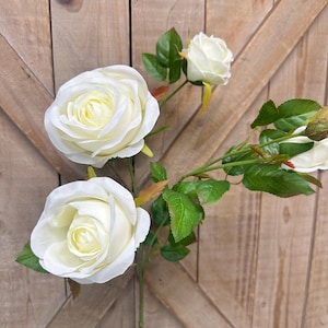 May include: A close-up of white artificial roses with green leaves against a wooden background. The roses are in various stages of bloom, with two fully open and one in bud. The petals are a creamy white color.