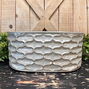 May include: A galvanized metal planter with a textured, repeating pattern. The rectangular container has a wide opening and is suitable for indoor or outdoor use. The planter is displayed against a wooden background.