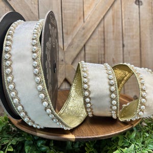 May include: Ivory velvet ribbon with a gold backing and pearl trim. The ribbon is rolled on a spool and displayed in a decorative arrangement. The ribbon is on a wooden tray with greenery.