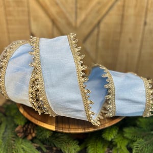 May include: A light blue decorative ribbon with gold trim and pearl accents. The ribbon is curled and rests on a wooden plate, surrounded by pine needles and a pine cone. The ribbon's edges are adorned with intricate gold lace.