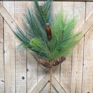 May include: A decorative pine branch with green needles and brown pine cones. The branch has a textured, gold-colored stem and is set against a wooden background. The arrangement is suitable for holiday or seasonal decor.